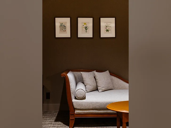 ベージュの長椅子、木製テーブル、植物画が飾られた部屋の一角